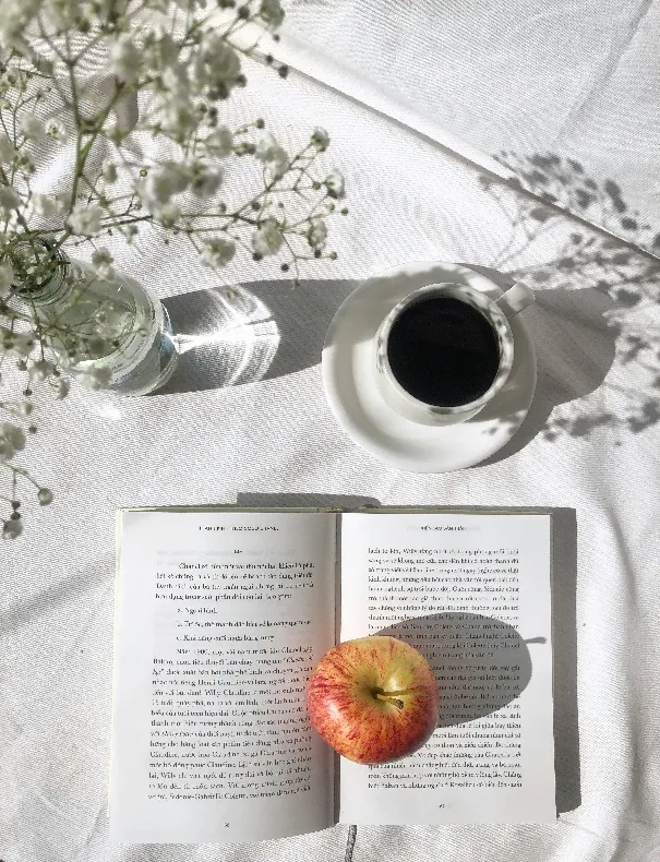A cozy aesthetic morning workspace with an open book, a cup of coffee, and a fresh red apple on a white table cloth in soft sunlight.