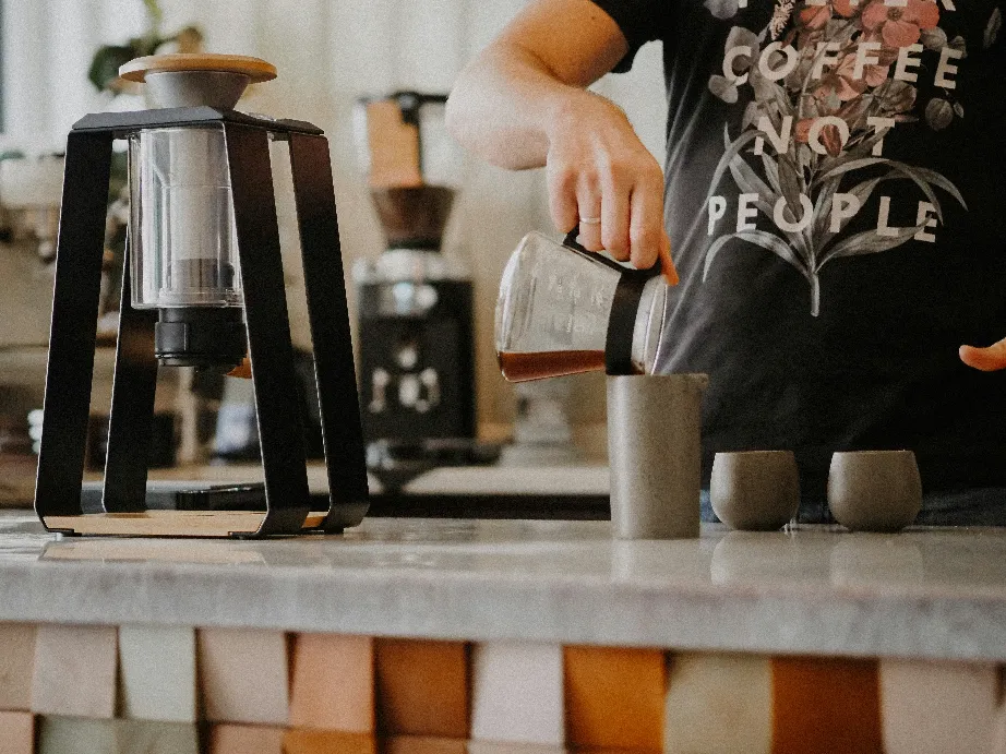 A barista pouring hot water from a kettle into a coffee dripper on a scale, brewing fresh pour-over coffee in a modern cafe.
