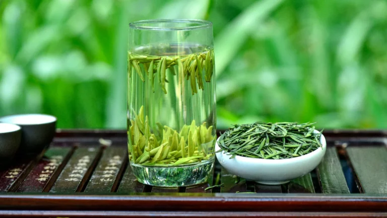 Fresh green tea leaves brewing in a tall clear glass of water with distinct vertical tea buds, next to a white ceramic dish filled with loose dry tea leaves on a wooden tea tray.