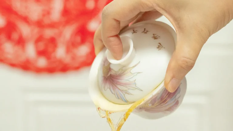 A close-up of a hand pouring freshly brewed tea from a white porcelain gaiwan with a floral pattern, demonstrating how to tilt the lid to filter the tea leaves.