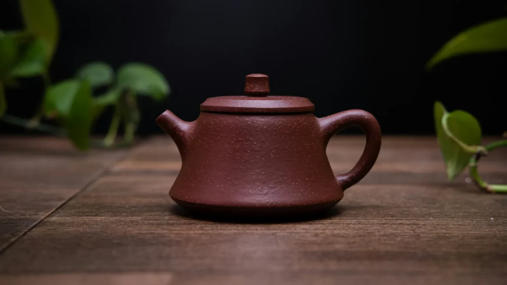 A close-up of a classic reddish-brown Yixing clay teapot sitting on a dark wooden table with a green plant in the blurred background.