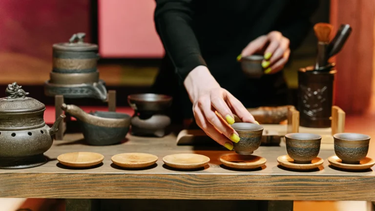 A person with green nail polish is pouring tea from a small, traditional dark earthenware Chinese gaiwan into tiny, dark-fired tea cups on a wooden tray, showing a gongfu tea brewing setup.