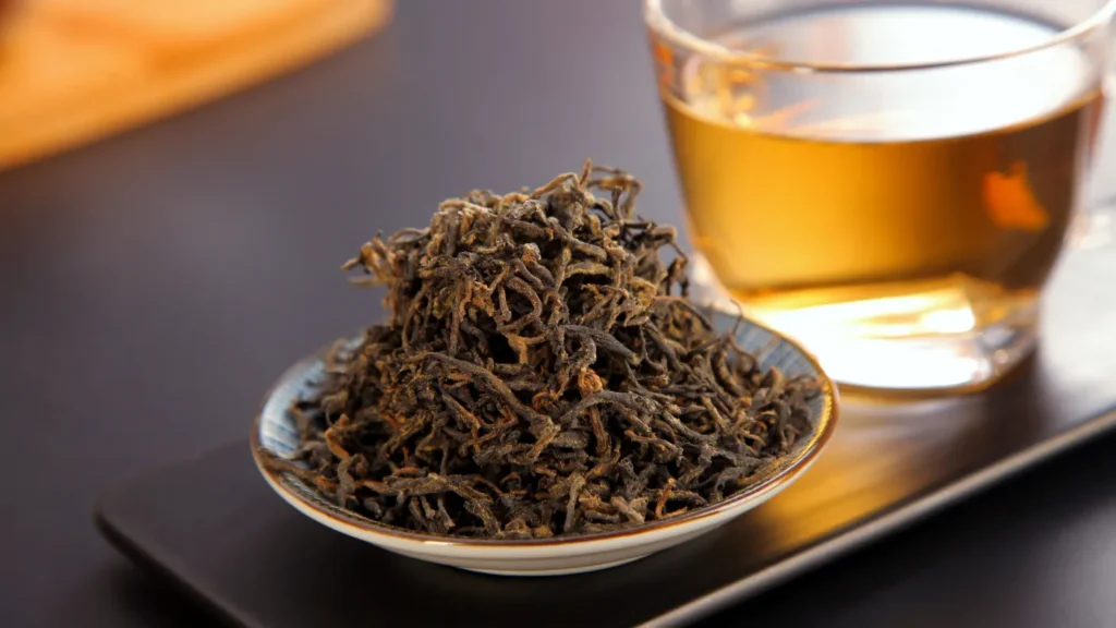 A bowl of dried Da Hong Pao tea leaves next to a glass cup of brewed oolong tea on a dark tray, illustrating Wuyi rock tea.