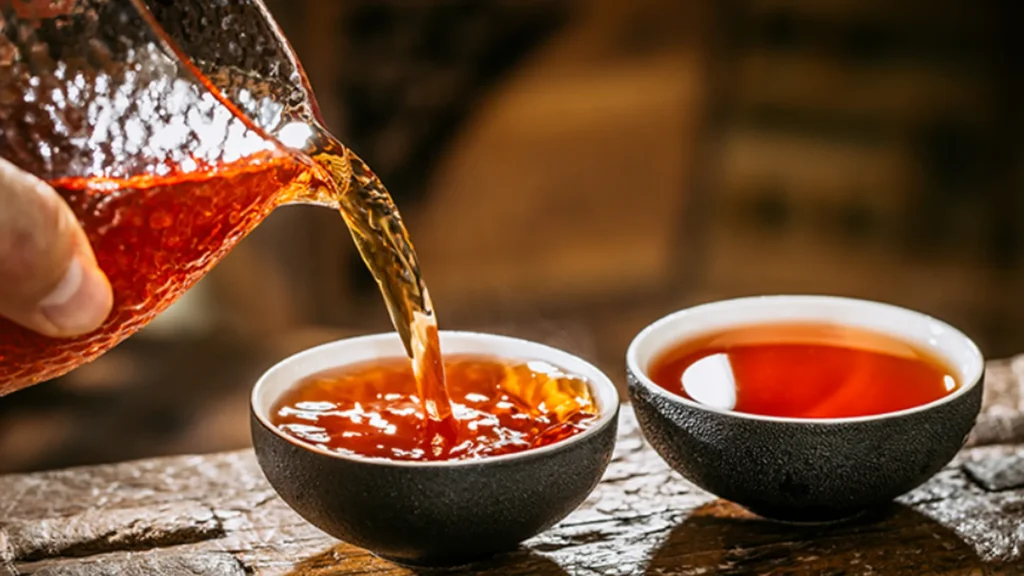 Freshly brewed Chinese black tea being poured from a glass pitcher into two small ceramic tasting cups on a wooden tea table.