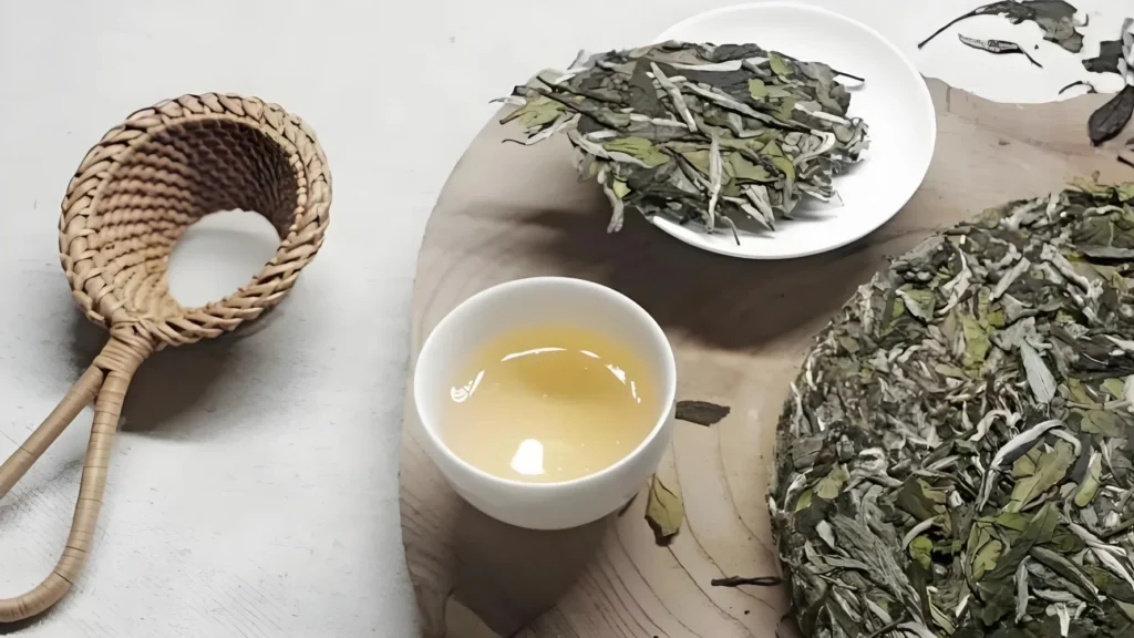 A loose leaf Chinese white tea setup featuring dried silver needle tea leaves in a wooden scoop next to a poured cup of pale yellow tea.